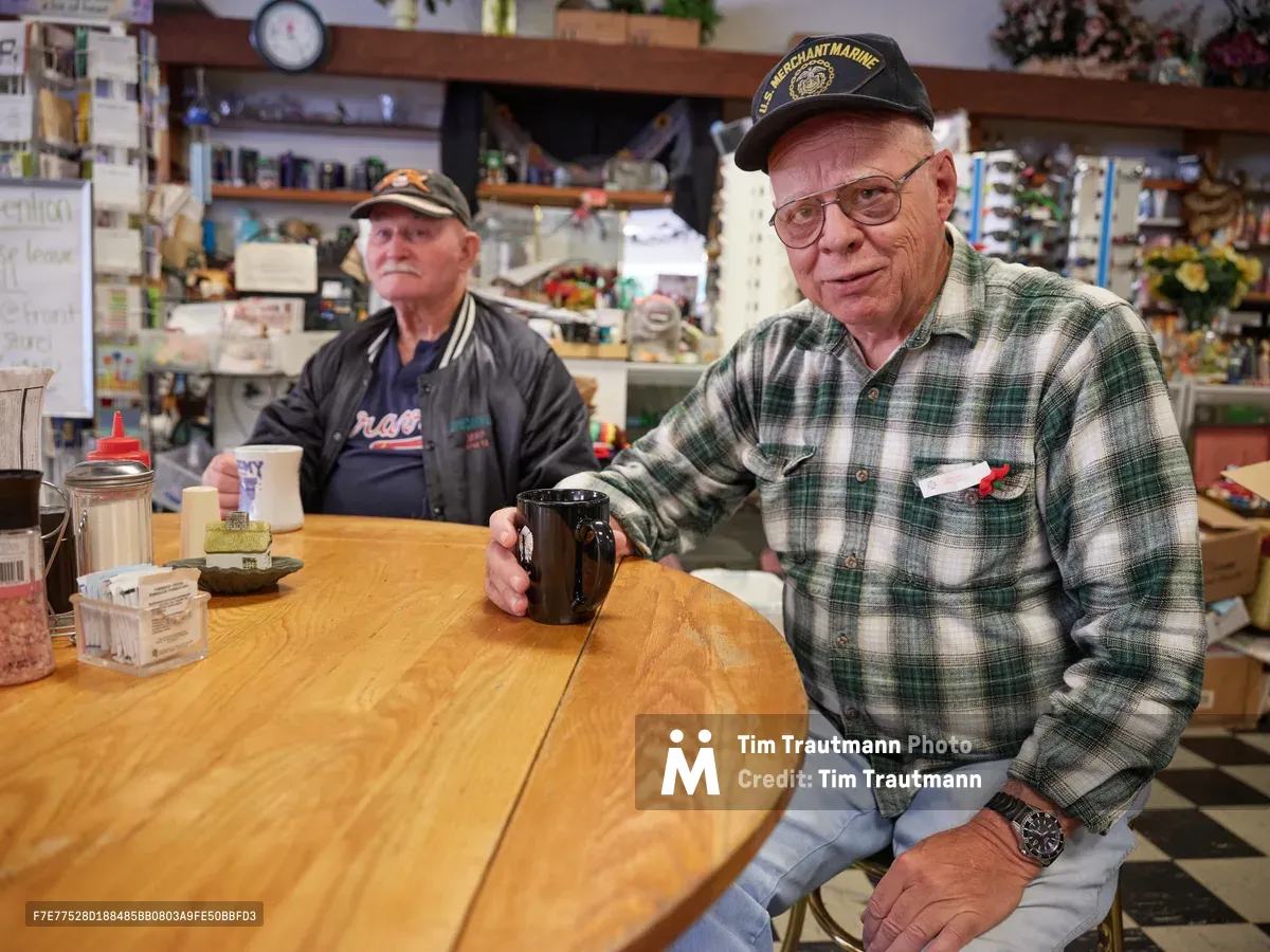Ron, a weathered merchant marine veteran in wire-rimmed glasses and plaid flannel, sits at a well-worn wooden table beside his longtime friend Jewel in this intimate portrait of St. Johns neighborhood camaraderie. The warm, diffused lighting illuminates decades of community history gathered around steaming coffee cups, while shelves lined with local memorabilia and ephemera create a backdrop rich with Portland's working-class heritage. Their relaxed postures and knowing expressions speak to countless mornings spent in this converted Ford dealership turned beloved neighborhood institution.