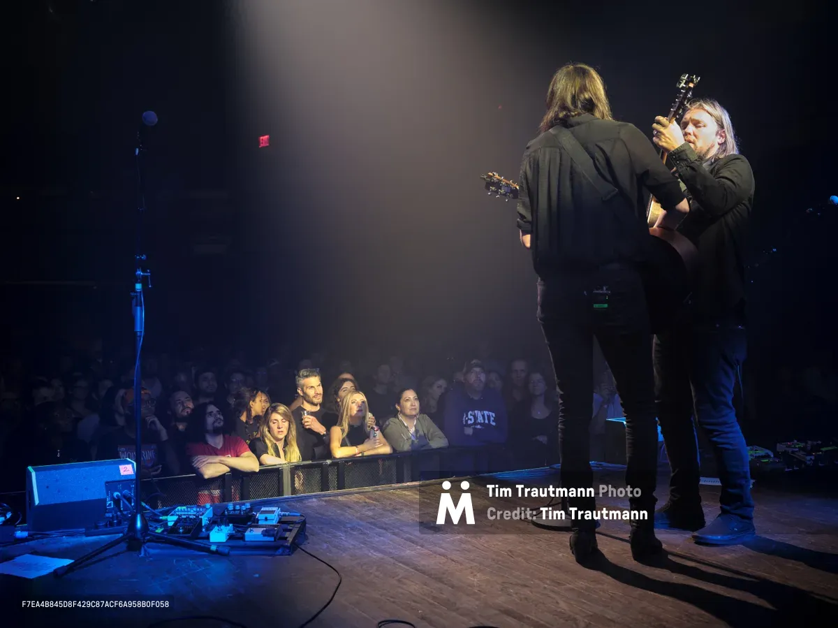 Emma Richardson and Russell Marsden of Band of Skulls perform during their 10 Year Anniversary Tour at Webster Hall in Manhattan's East Village. The duo stands center stage bathed in dramatic stage lighting, with Richardson holding her guitar while Marsden faces away from the camera. The intimate venue setting captures the engaged audience seated at tables in the foreground, creating a sense of connection between performers and fans in this historic New York music venue.