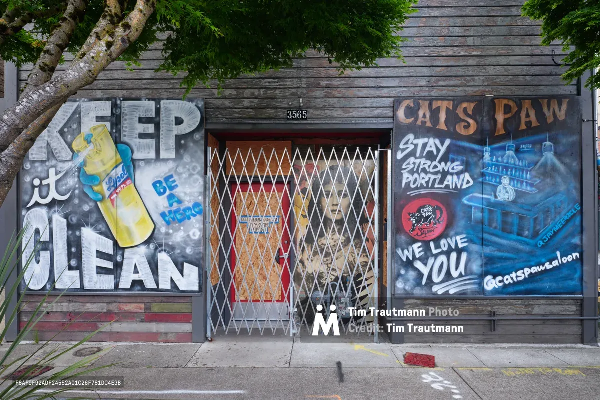 Weathered wooden storefronts along Southeast Division Street display contrasting pandemic-era murals beneath overhanging maple branches. The left mural promotes hygiene with "KEEP it CLEAN" messaging and sanitizer imagery, while the right celebrates local resilience with "CAT'S PAW Stay Strong Portland We Love You" in bold lettering. A security grate protects the central entrance at address 3565, creating an urban tableau of community solidarity and public health awareness.