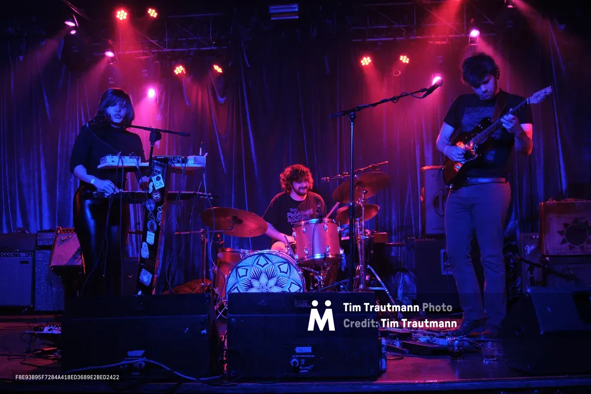 Experimental band Sun Blood Stories commands the intimate stage at Dante's in downtown Portland, bathed in crimson and violet stage lights that pierce through the smoky atmosphere. The three-piece band from Boise, Idaho—featuring a keyboardist, drummer, and guitarist—creates their mesmerizing modern-day opera sound amid the venue's signature industrial backdrop of exposed metal rigging and curtained walls. The moody lighting casts dramatic shadows while highlighting the concentrated energy of the musicians mid-performance.