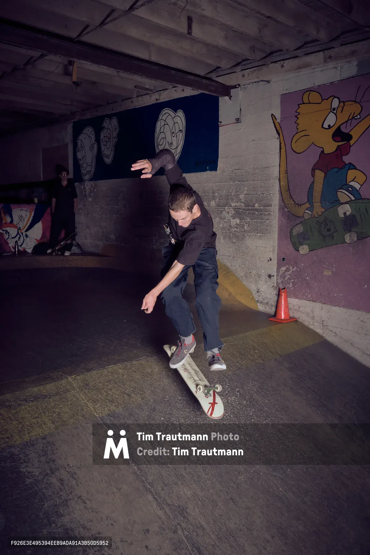 A skateboarder executes a trick in the dimly lit basement of Commonwealth Skateboarding in Portland's Buckman neighborhood. The concrete space is adorned with colorful cartoon murals, including character faces on blue backgrounds and a Simpsons-style figure. Flash photography captures the raw energy of the underground skate scene, with another figure visible in the shadowy background as wooden ceiling beams frame the gritty urban sanctuary.