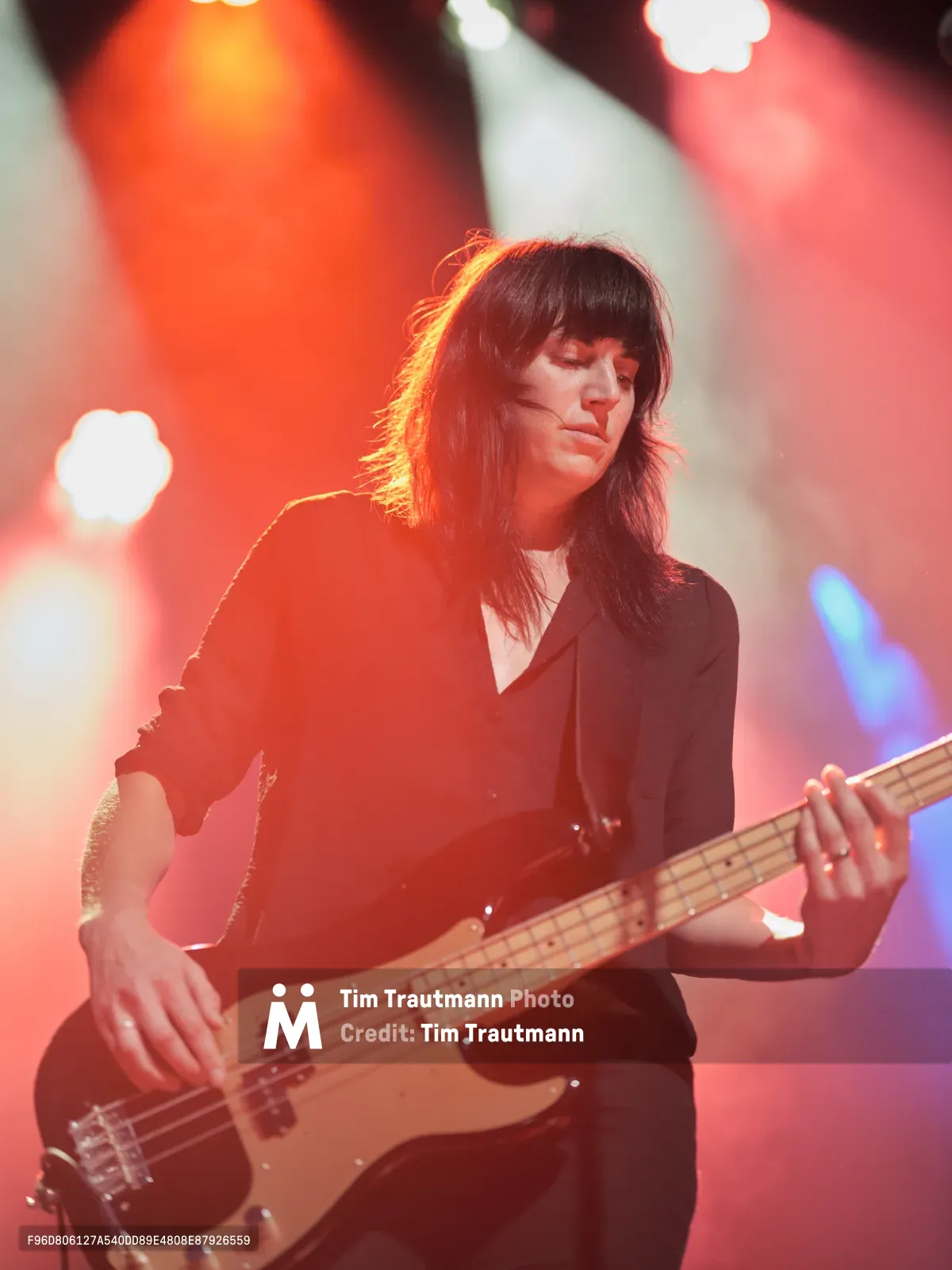 Emma Richardson of Band of Skulls commands the stage at Webster Hall during the band's 10 Year Anniversary Tour, her bass guitar gleaming under theatrical lighting. Dramatic amber and red stage lights create a moody atmosphere, casting her dark hair and black attire in silhouette while highlighting her focused expression. The intimate Manhattan venue's iconic lighting rig creates bokeh effects in the background, emphasizing the raw energy of live performance.