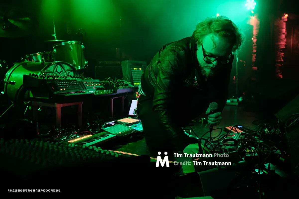 Electronic musician Eli Savage, performing as Ellipsism, leans intently over his synthesizer setup during a live performance at RXFest 2019. Bathed in vivid emerald stage lighting that transforms the intimate venue into an otherworldly sonic laboratory, the bearded artist manipulates a dense array of electronic equipment while gripping a microphone. The atmospheric lighting casts deep shadows across Dante's club in Portland's downtown district, creating a moody tableau where technology and artistry converge in the underground music scene.