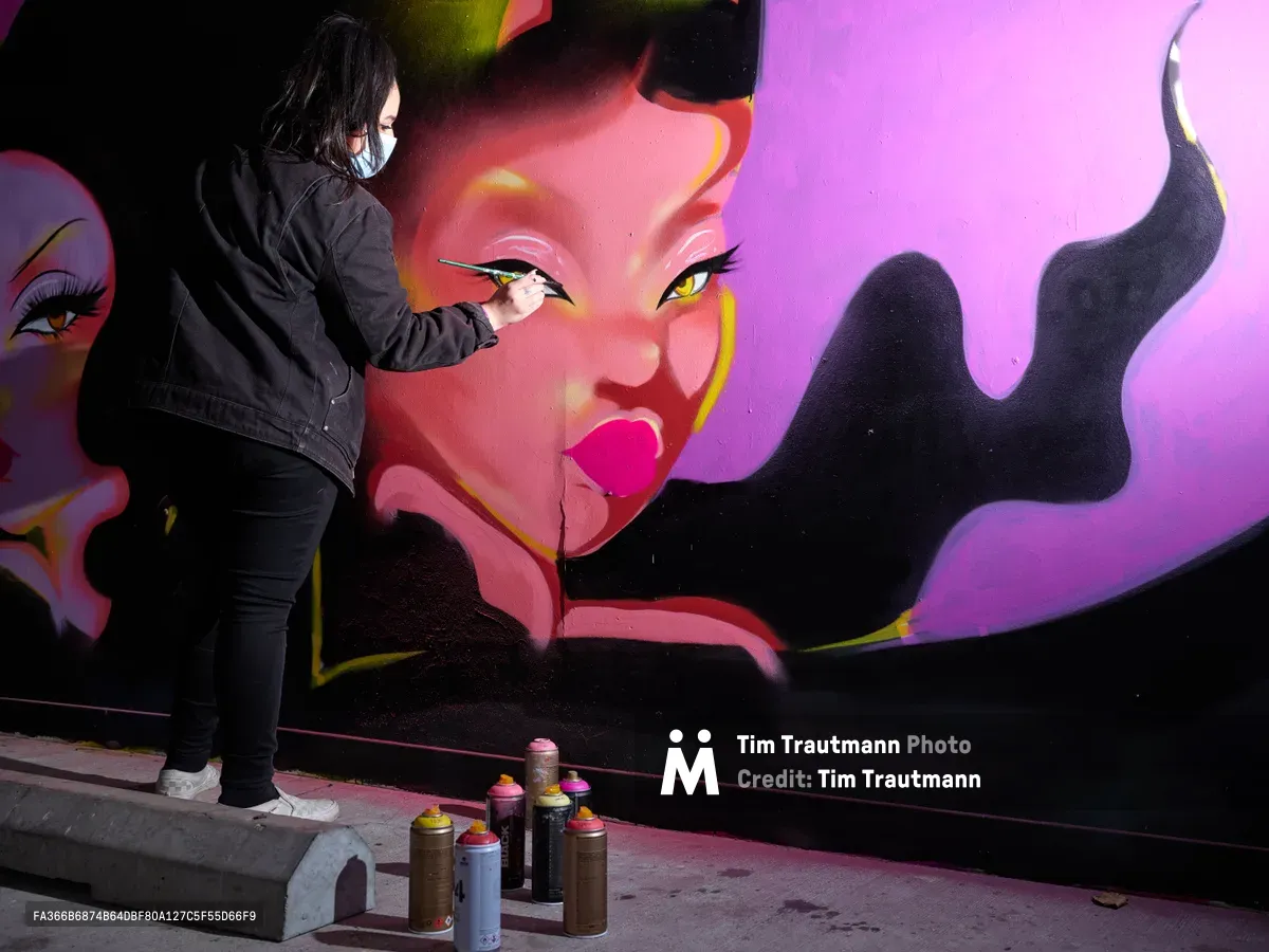 Multidisciplinary artist Vilatose meticulously applies paint to a striking feminine portrait rendered in electric pinks and purples against the concrete wall of Clay Creative's parking garage in Portland's Hosford-Abernethy neighborhood. The masked artist, clad in all black, works with focused precision as an arsenal of spray paint cans sits ready on the pavement below. The mural depicts a sultry female figure with dramatic eyeliner and flowing dark hair, her image emerging from shadows into brilliant magenta light.