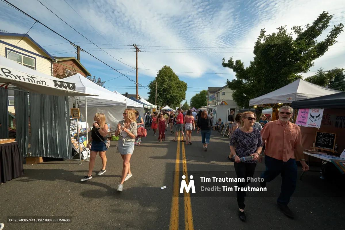 Vendors and visitors gather along the closed street during Portland's annual Mississippi Street Fair, with white pop-up tents lining both sides of the asphalt roadway. A mature couple in casual summer attire walks toward the camera in the foreground, while dozens of festival-goers browse vendor stalls beneath a partly cloudy afternoon sky. The residential neighborhood's modest homes and power lines frame this quintessential Pacific Northwest community event.