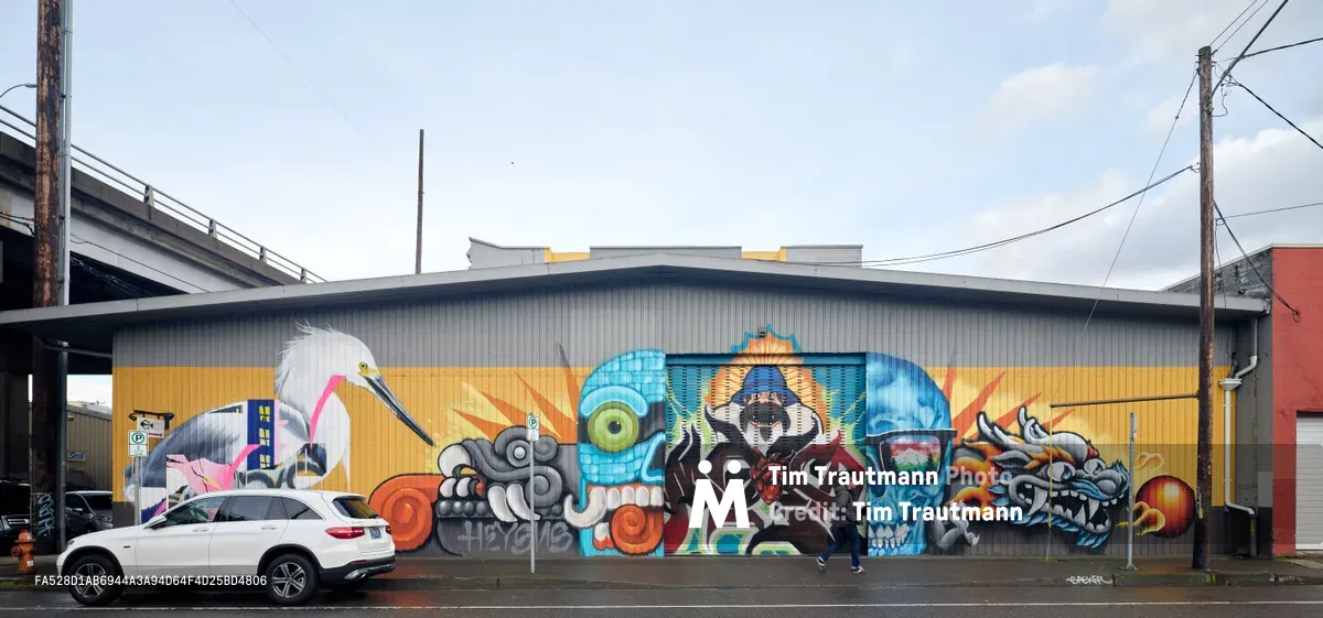 A vibrant warehouse wall mural sprawls across an industrial building in Portland's Central Eastside, showcasing a fantastical collection of characters including a white heron, mechanical creatures, skulls, and mythological figures rendered in electric blues, fiery oranges, and golden yellows. A white SUV sits parked against the colorful corrugated metal canvas, while power lines and an overpass frame the urban art installation under an overcast Pacific Northwest sky. The commissioned artwork transforms the utilitarian industrial landscape into a dynamic visual narrative.