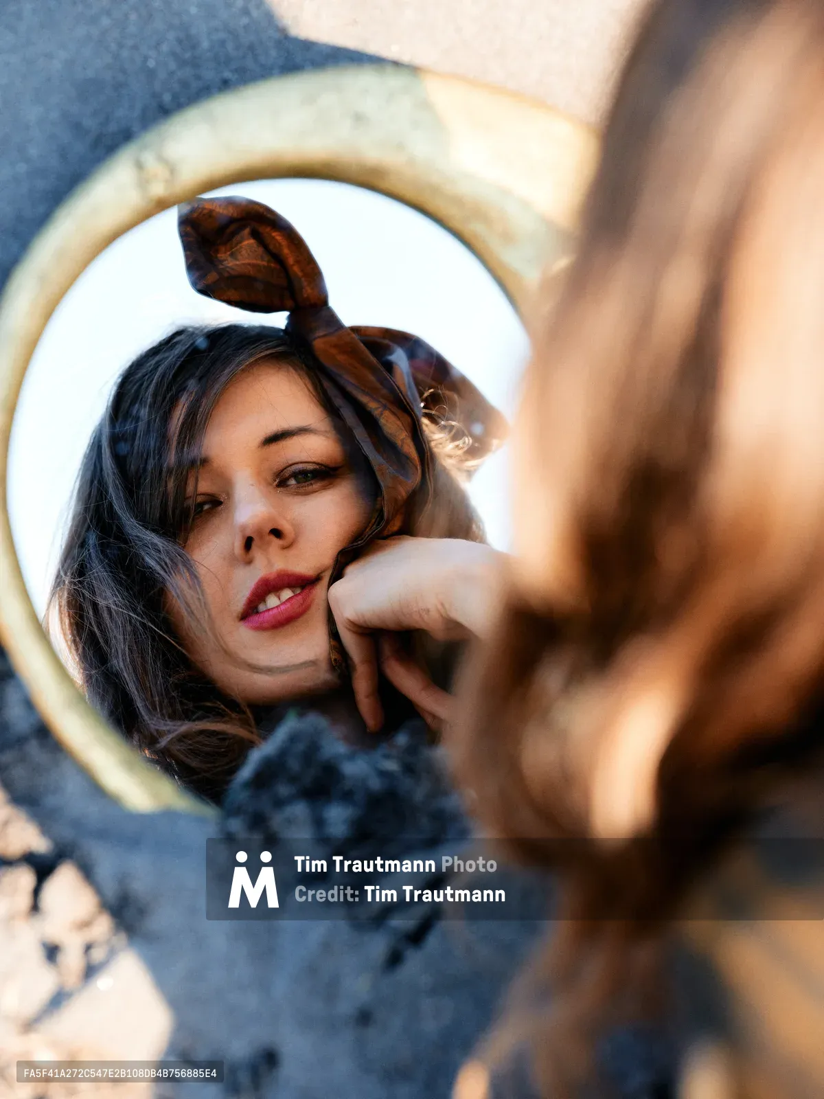 A contemplative moment captured through a vintage hand mirror held along the Floodplain Trail in Multnomah County, Oregon. The reflection reveals a young woman with dark hair adorned with a tortoiseshell headband, her red lips and gentle expression framed by the mirror's weathered brass edge. Warm golden sunlight filters through the scene, creating an intimate interplay between the reflected subject and the blurred riverbed environment surrounding the trail.