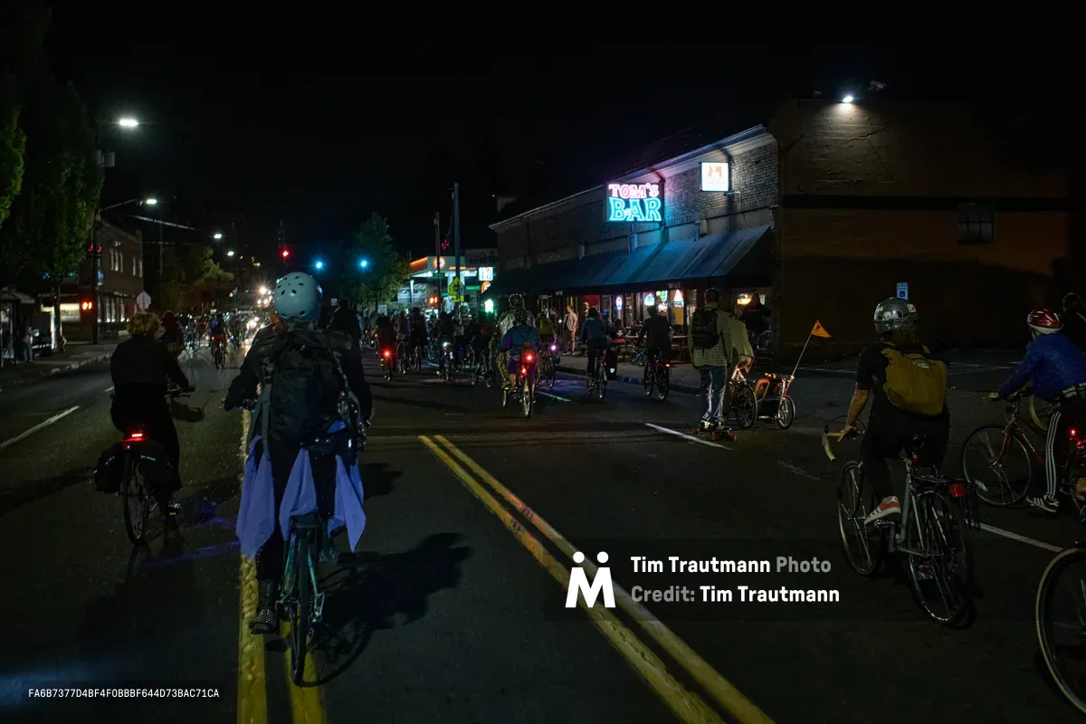 Dozens of cyclists converge on Southeast Division Street during Portland's Drop Out Prom Ride, part of the city's annual Pedalpalooza celebration. The nocturnal scene pulses with red bicycle lights scattered across the asphalt like urban fireflies, while Tom's Bar's neon sign casts an electric blue glow over the assembled riders. Helmeted participants in casual attire cluster in the street and on sidewalks, their bicycles creating an impromptu street festival atmosphere beneath the warm amber streetlights of this Richmond neighborhood intersection.