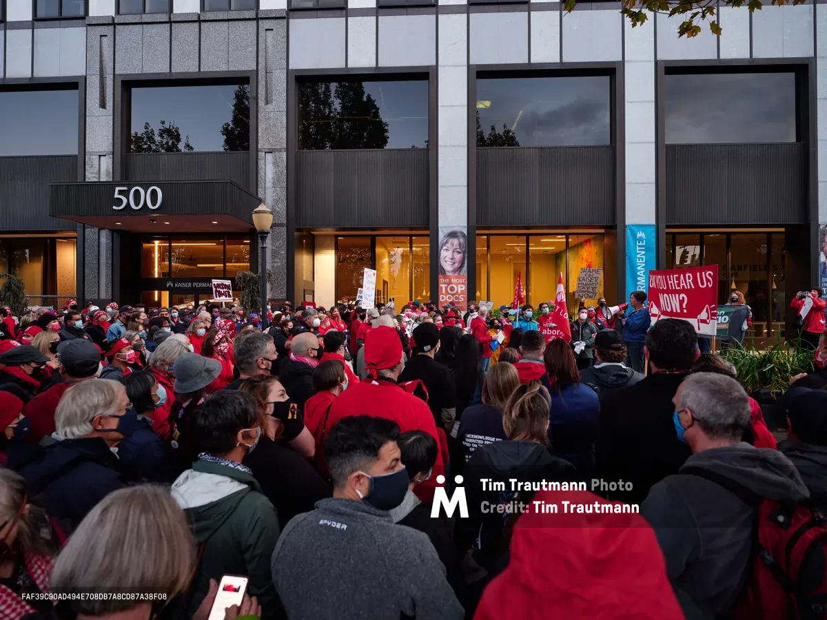 A dense crowd of healthcare workers, predominantly wearing red jackets and face masks, congregates outside the Kaiser Permanente Tower at 500 Northeast Multnomah Street in Portland's Lloyd District. The sea of protesters fills the frame from foreground to the building's glass facade, creating a powerful visual of collective action. Protest signs punctuate the crowd, including one reading 'CAN YOU HEAR US NOW?' while the building's modernist architecture looms behind them, its illuminated interior contrasting with the gathering dusk outside.