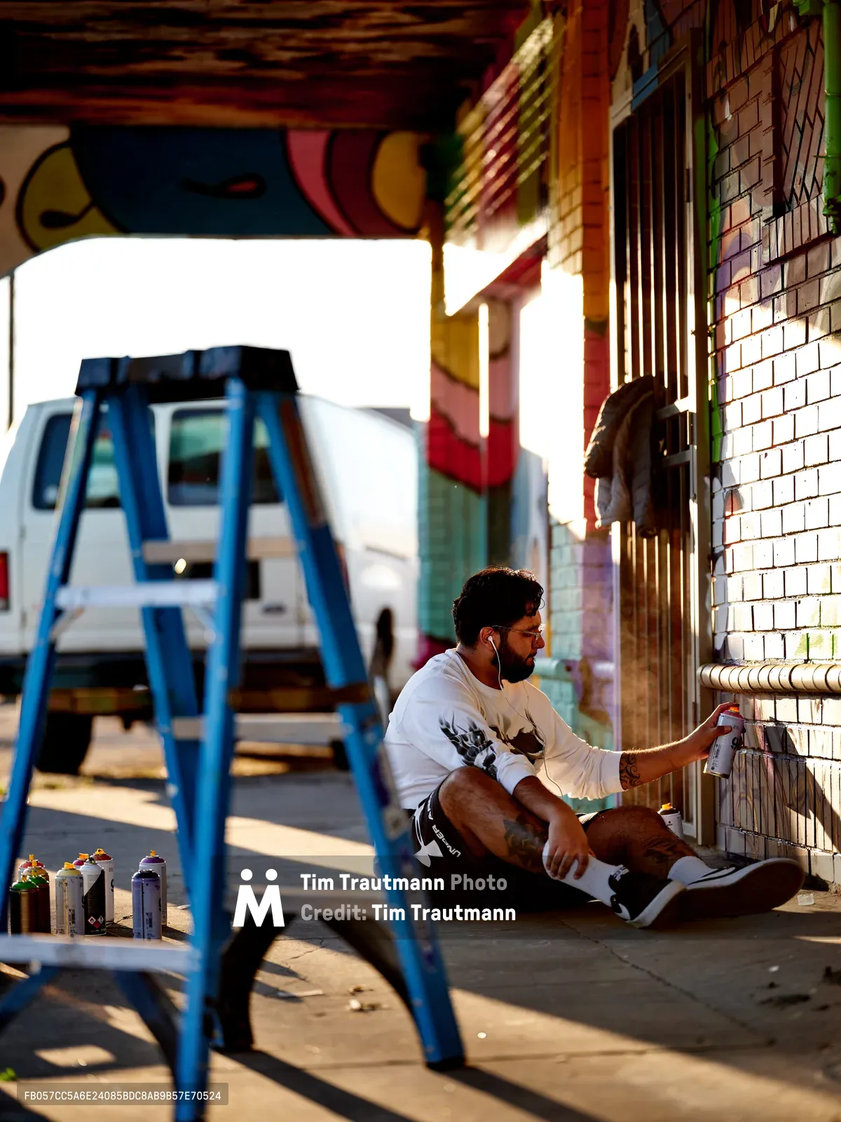 Muralist Heysus sits cross-legged on a sidewalk in Portland's Alberta district, spray painting intricate details on a colorful brick wall mural. Golden hour light streams through the urban scene, casting warm shadows from a blue metal stepladder in the foreground while a white RV sits parked in the soft-focus background. The artist works with focused concentration, earbuds in, surrounded by scattered spray paint cans on the concrete.