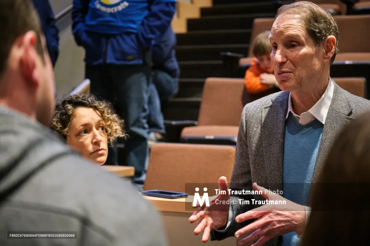 Senator Ron Wyden leans forward in animated conversation during a town hall meeting in Portland's Saint Johns neighborhood, his expressive hands gesturing as he addresses concerns from local constituents. The intimate setting captures the democratic process in action, with warm wooden auditorium seating creating a backdrop for civic engagement. A woman with curly hair listens intently nearby, while the senator's blue sweater and herringbone jacket reflect the casual yet professional tone of the community gathering.