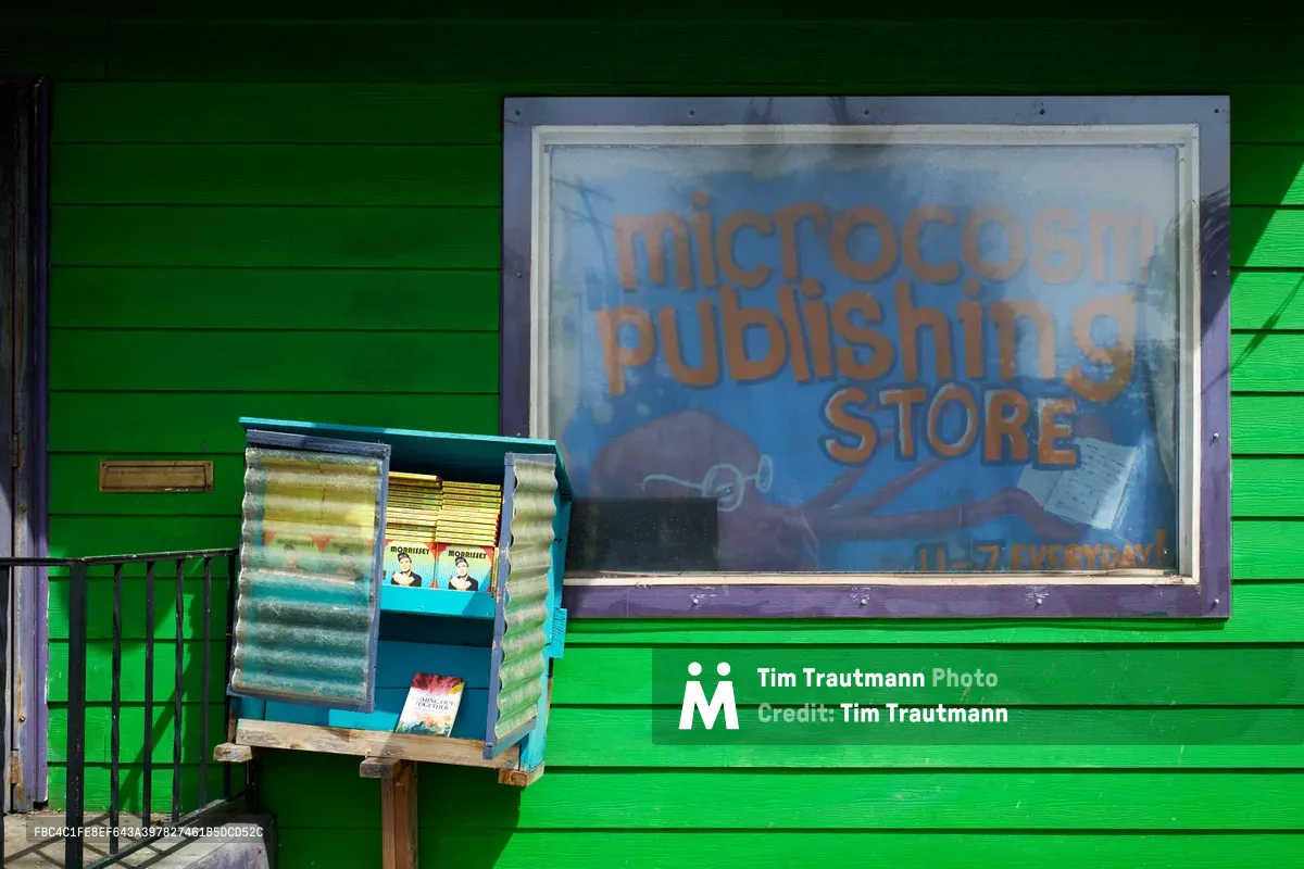 A small free library box sits beneath the faded signage of Microcosm Publishing Store on North Williams Avenue in Portland's historic Eliot neighborhood. The community book exchange stands against the building's vibrant green clapboard siding, while the store's blue window display promises "11-7 EVERYDAY" hours. Afternoon shadows cast geometric patterns across this neighborhood literacy resource at this grassroots literary outpost.