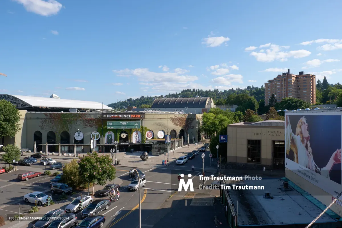 An elevated view captures the bustling urban landscape surrounding Providence Park stadium in Portland's Goose Hollow neighborhood. The distinctive curved architecture of the soccer venue dominates the center frame, while residential high-rises and forested hills create a layered backdrop under partly cloudy skies. Street-level activity unfolds below with parked vehicles and pedestrians navigating the intersection, while a large billboard advertisement anchors the right side of the composition.