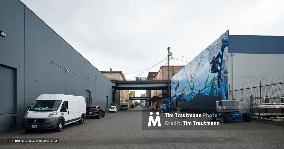 Artists Jeremy Nichols and Devin Finley work from a blue aerial lift to create a large-scale oceanic mural on the side of an industrial building in Southeast Portland's Central Eastside district. The overcast sky creates even lighting across the industrial scene, where gray warehouse buildings frame the street and an overpass connects the urban landscape. A white work van and safety equipment scattered on the asphalt emphasize the active nature of this public art installation commissioned by the Portland Street Art Alliance.