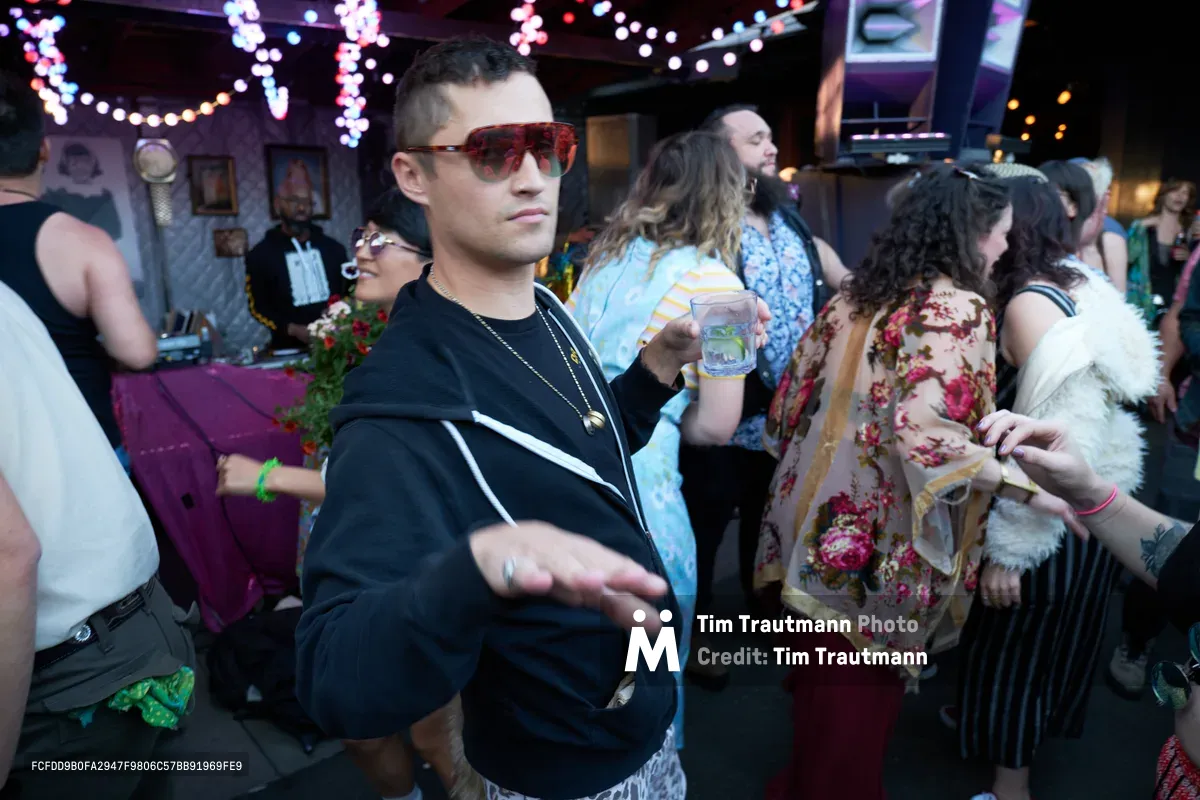 A man in tortoiseshell aviators and a black tracksuit gestures mid-conversation while holding a drink at Your Sunday Best, a vibrant summer day party at Portland's White Owl Social Club. The intimate venue pulses with string lights and eclectic wall art as diverse partygoers in floral prints and casual attire mingle in the warm, bokeh-lit atmosphere. The scene captures the relaxed yet energetic spirit of Portland's underground music scene, where afternoon revelry unfolds beneath cascading fairy lights.