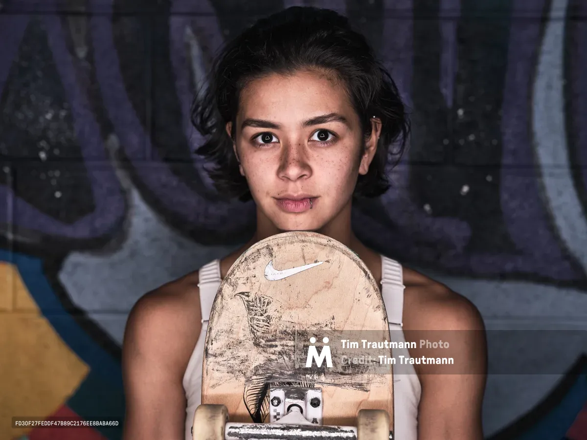 A close-up portrait of pro skateboarder Christiana Means holding a well-worn Nike skateboard deck up to her chin, gazing directly into the camera with a calm, focused expression. She has short dark hair, freckles, and a small lip piercing, and wears a white tank top. A colorful graffiti mural in purple, blue, and yellow serves as the background.