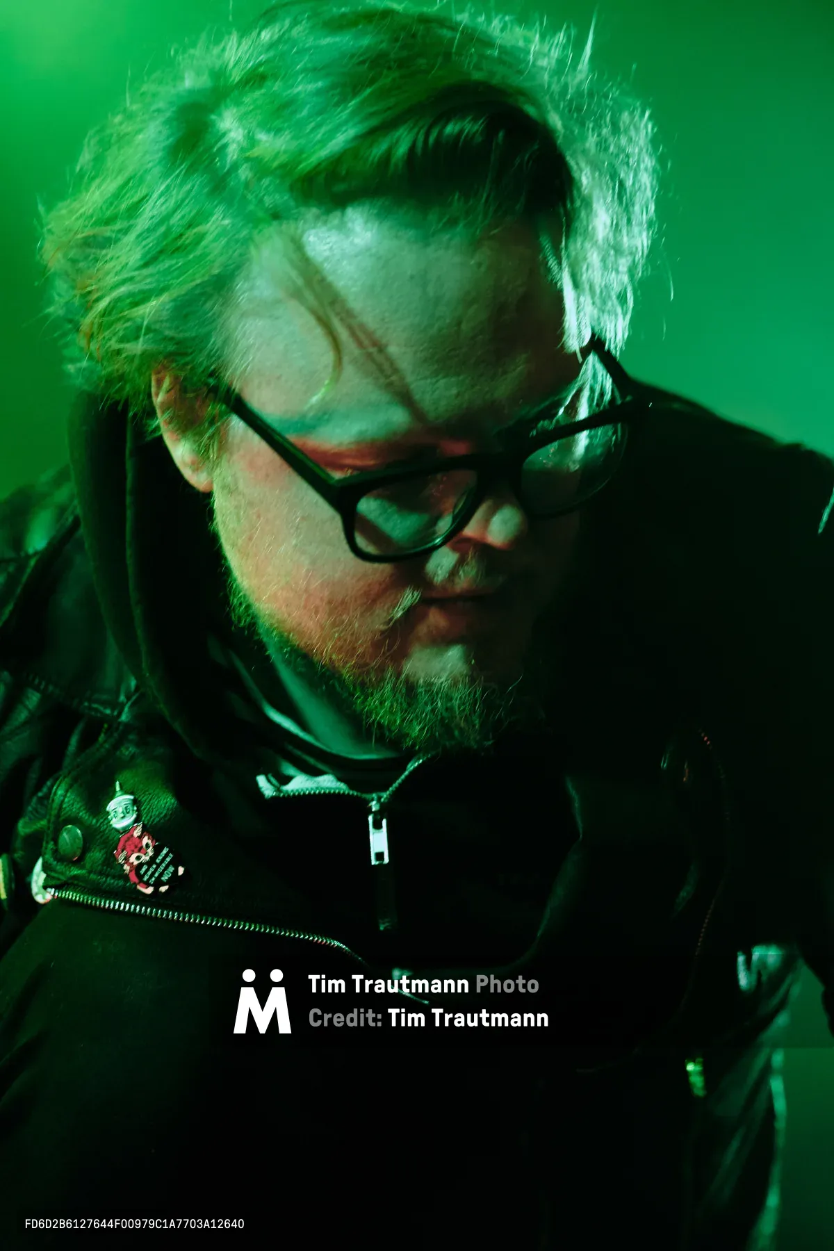 Electronic musician Eli Savage, performing as Ellipsism, becomes lost in concentration during his live set at RXFest 2019. Bathed in atmospheric green lighting at Dante's venue in Portland, Oregon, his silver hair catches the theatrical glow as he manipulates his equipment. The intimate portrait captures the artist's complete immersion in his craft, black-rimmed glasses reflecting the stage lights while patches on his dark jacket hint at his underground aesthetic.