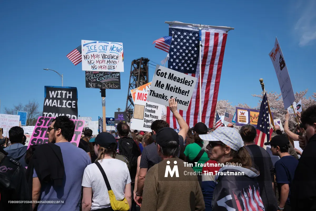 Demonstrators gather beneath the clear blue Oregon sky on Northwest Naito Parkway in Portland's historic Old Town district, their voices rising in unified dissent. American flags flutter alongside handmade signs declaring messages against government overreach, including prominent placards reading "ICE OUT NOW!" and "Got Measles? Me Neither!" The crowd stretches into the distance, a sea of diverse faces and colorful protest signs framed by the industrial architecture of Portland's waterfront. Spring light illuminates the passionate assembly as citizens exercise their constitutional right to peaceful protest.