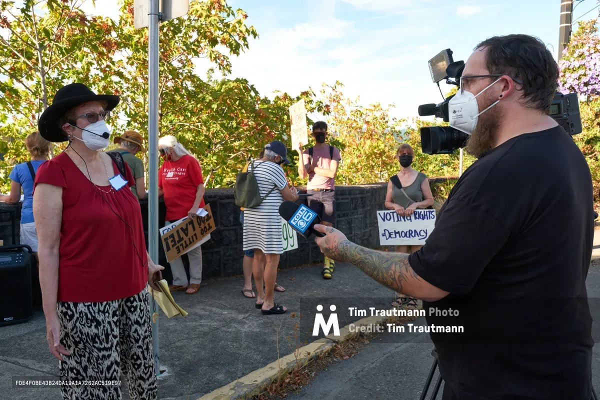 A tattooed television cameraman from KOIN Channel 6 interviews a climate activist wearing a red top and black hat outside Representative Kurt Schrader's Oregon City office. The woman stands confidently before the camera while fellow demonstrators gather behind her, their handmade signs advocating for voting rights and democracy visible in the dappled afternoon light. The scene unfolds on McLoughlin Promenade, where autumn foliage frames this grassroots moment of civic engagement.
