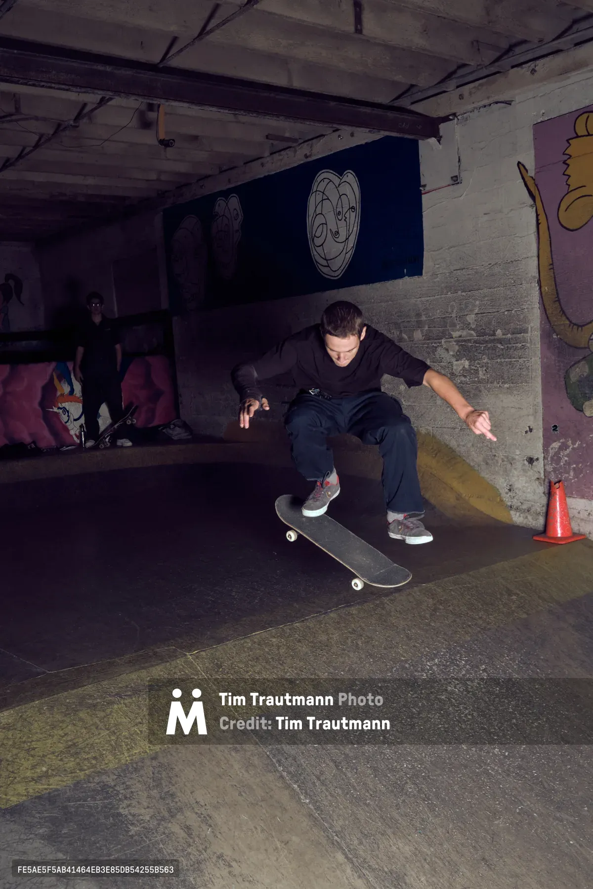 A skateboarder navigates the dimly lit basement of Commonwealth Skateboarding in Portland's Buckman neighborhood, his form silhouetted against vibrant graffiti murals. The industrial concrete space, marked by exposed ceiling beams and painted brick walls, creates an intimate underground atmosphere where street art and skateboarding culture converge. Harsh artificial lighting casts dramatic shadows as the athlete maintains balance mid-trick, while a solitary traffic cone punctuates the raw urban setting.