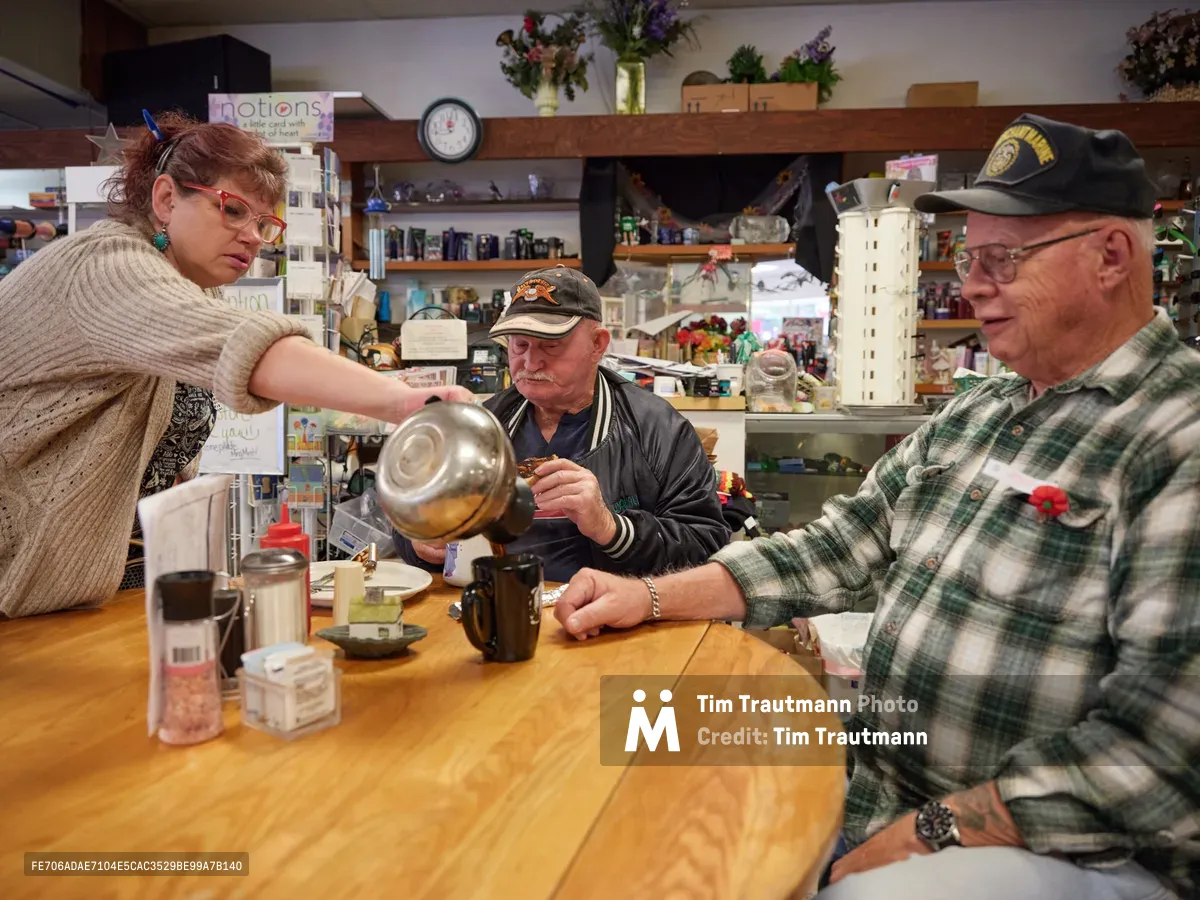 Inside the cozy confines of Pattie's Home Plate Café in Saint Johns, Portland, a woman in a cream cable-knit sweater pours steaming coffee from a glass carafe for two elderly patrons seated at a worn wooden counter. The men, one wearing a baseball cap and the other in a green plaid flannel shirt, wait patiently as warm light illuminates the cluttered, lived-in interior filled with shelves of collectibles and everyday café paraphernalia. The scene captures the intimate ritual of neighborhood hospitality in this beloved Portland community gathering place.