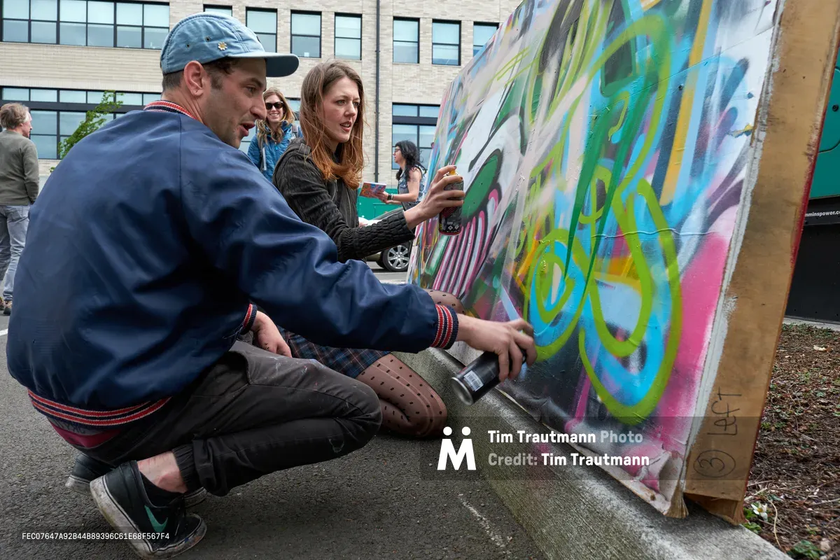 At the Taylor Electric Blocks in Portland's Hosford-Abernethy neighborhood, two artists crouch beside a vibrant mural in progress, spray cans in hand as they add fresh layers of green, blue, and pink aerosol paint to the collaborative canvas. The scene captures the intimate, concentrated energy of live street art creation, with onlookers gathered in the background beneath the industrial brick facade of the surrounding buildings. The demonstration unfolds on concrete, where the temporary wooden panel serves as both easel and public art platform.
