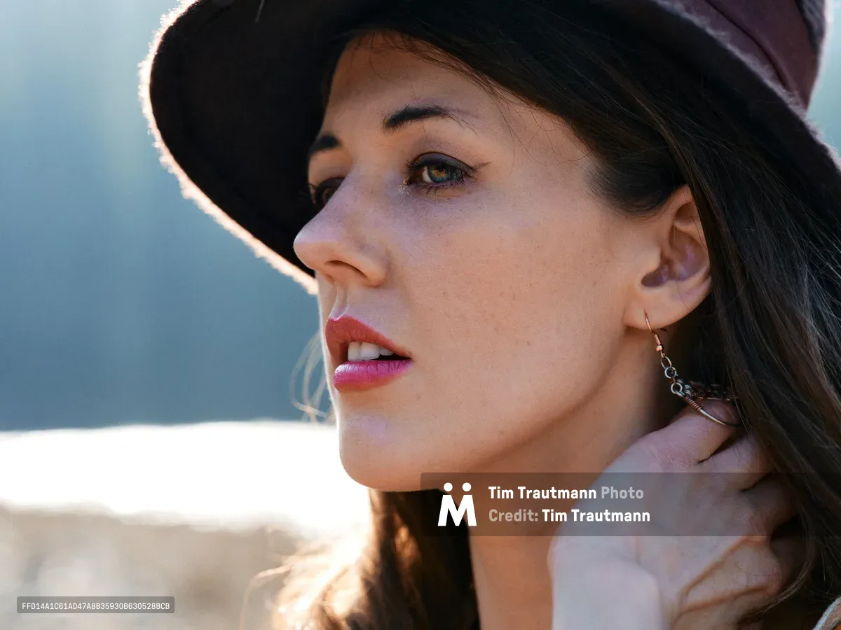 A young woman gazes contemplatively into the distance, her profile illuminated by warm golden light filtering through her wide-brimmed hat. The intimate portrait captures her wearing delicate chain earrings and a soft pink garment, with the dreamy bokeh of Oregon's riverbed trail creating an ethereal backdrop. The gentle lighting emphasizes the natural texture of her skin and the subtle gradation from shadow to light across her features.