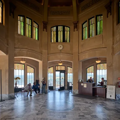 The grand interior of Vista House at Crown Point reveals its majestic Art Nouveau architecture, with soaring limestone walls and distinctive stained glass windows filtering golden afternoon light. Visitors gather beneath the ornate two-story rotunda, some seated on benches while others explore the information desk, creating an intimate human scale against the monument's imposing classical proportions. The polished marble floors reflect the warm illumination streaming through the geometric leaded glass panels, while a vintage clock marks time in this beloved Oregon landmark overlooking the Columbia River Gorge.