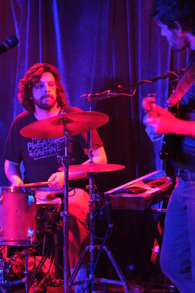A curly-haired drummer from Sun Blood Stories commands the kit at Portland's legendary Dante's venue, his form bathed in intense red and purple stage lighting. The atmospheric illumination transforms the intimate club setting into a moody sonic sanctuary, with vertical light beams cutting through the haze behind him. His bandmate's silhouette lurks in the background shadows, creating depth in this electrifying live performance moment.