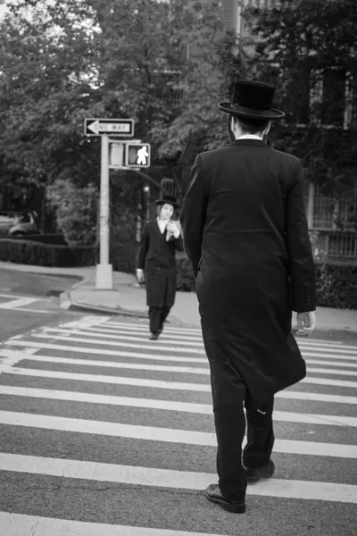 A black and white street photograph captures an intimate moment between two Orthodox Jewish males crossing a zebra-striped crosswalk in Williamsburg, Brooklyn. The foreground figure, wearing a traditional black coat and hat, dominates the frame with his back to the camera, while a younger person approaches from the opposite direction near street signage. Dappled sunlight filters through leafy trees, creating atmospheric contrast against the urban streetscape of this historic neighborhood.