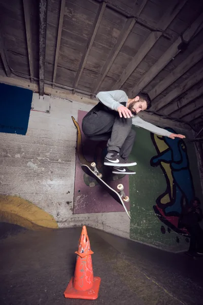 A skateboarder poses confidently in the raw industrial interior of Commonwealth Skateboarding in Portland's Buckman neighborhood. The young man sits casually against weathered concrete steps beneath exposed wooden ceiling beams, his skateboard resting nearby alongside a bright orange traffic cone. Moody lighting illuminates the gritty urban architecture, capturing the authentic underground skateboarding culture that defines Portland's Southeast 20th Avenue scene.
