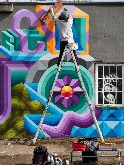 A muralist in gray hoodie and beanie balances precariously on a stepladder, adding finishing touches to a vibrant geometric wall painting in Northeast Portland, Oregon. The kaleidoscopic mural features bold dimensional typography and mandala-like patterns in electric purples, teals, oranges, and blues against weathered gray concrete. Spray paint cans and art supplies scattered below reveal the creative process, while diffused daylight illuminates the transformation of urban space into colorful public art.