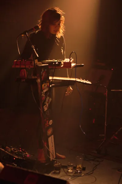 A female musician from experimental band Sun Blood Stories performs on keyboard at Dante's venue in downtown Portland, her silhouette dramatically lit by warm amber stage lighting that creates a halo effect around her tousled hair. She stands focused behind a multi-tiered keyboard setup, surrounded by the intimate atmosphere of the underground music scene. The moody lighting and shadowy backdrop capture the raw energy of this Boise-founded band's mesmerizing live performance.