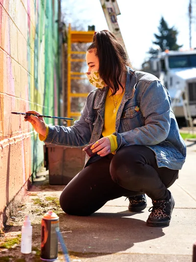 Multidisciplinary artist Vilatose crouches beside a weathered brick wall on Northeast Alberta Street, delicately applying paint to her emerging mural titled Jardín Cubana, which depicts three female figures. Golden afternoon light illuminates her yellow sweater and denim jacket as she works with focused concentration, her patterned face mask a contemporary reminder of the times. Spray paint cans rest on the concrete sidewalk beside her, while the colorful urban landscape of Portland's Alberta Arts District provides a vibrant backdrop to this moment of creative expression.