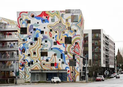A striking apartment building in Portland's Buckman neighborhood showcases an extraordinary psychedelic mural that completely envelops its facade. The massive artwork features flowing ribbons of vibrant blues, reds, yellows, and earth tones that cascade across the white surface, punctuated by geometric black squares positioned over windows. The overcast Pacific Northwest sky creates a muted backdrop for this explosion of color, while modern residential buildings and bare winter trees frame the scene along the quiet urban street.