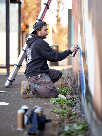 Travis Fields, known as campographic, kneels on the sidewalk applying spray paint to a mural of poet Amanda Gorman at the intersection of Northeast 15th Avenue and Killingsworth Street in Portland's Alberta neighborhood. The artist works in profile, his dark hoodie and backwards cap silhouetted against the warm afternoon light that bathes the urban streetscape. Paint cans scattered at his feet and a ladder positioned nearby speak to the methodical process of transforming this corner wall into public art.