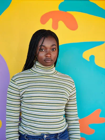 Naomi Likayi stands confidently before her completed mural in Portland's Alberta Arts District, her long braids framing her face as she gazes directly at the camera. She wears a vintage-inspired striped turtleneck in earth tones that harmonizes beautifully with the explosive colors behind her—brilliant turquoise, sunny yellow, coral orange, and purple organic shapes that seem to dance across the wall. The soft natural lighting creates an intimate yet powerful portrait that captures both the artist's quiet strength and the exuberant energy of her public artwork in this historically significant neighborhood.