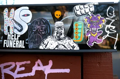A collection of provocative stickers and street art adorns a black window frame on North Mississippi Avenue in Portland, creating a visual collage of contemporary political dissent. The eclectic mix includes cartoon characters, political imagery, and countercultural symbols plastered against the urban storefront, with additional pink graffiti marking the rust-colored wall below. The juxtaposition of playful imagery with darker political commentary captures the raw energy of Portland's street art scene in this gentrifying neighborhood.