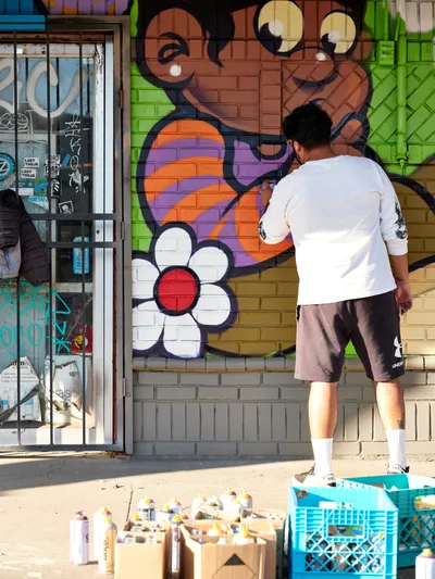 Muralist Heysus works intently on a colorful character mural featuring a cartoonish face with googly eyes and a simple white flower at the intersection of Killingsworth and NE 15th Avenue in Portland's Alberta district. Dressed casually in a white t-shirt and brown shorts, the artist applies spray paint to the brick wall while surrounded by crates of aerosol cans scattered across the concrete sidewalk. The afternoon light illuminates the vibrant oranges, purples, and greens of the emerging artwork against the urban storefront backdrop.