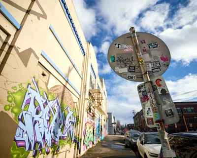 A vibrant graffiti mural dominates the cream-colored wall of a former Salvation Army warehouse in Portland's Central Eastside district, where purple and green wildstyle lettering creates a bold contrast against the weathered brick. A sticker-bombed street sign stands sentinel in the foreground, its circular surface layered with underground art collective logos and subcultural markers. The late afternoon light bathes the urban scene in warm tones while dramatic clouds sweep across the Pacific Northwest sky, creating an atmospheric backdrop to this commissioned street art installation.