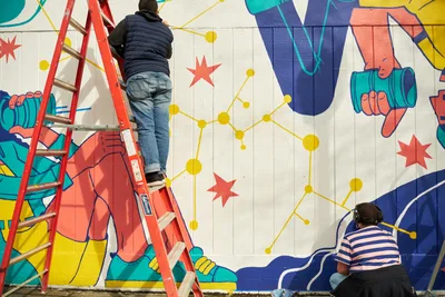 Two artists collaborate on an expansive public mural titled 'Let's Talk' on the exterior wall of Open Signal in Portland's Eliot neighborhood. One artist works from a red ladder, carefully painting details on the white concrete block surface, while another artist in a striped shirt works at ground level with headphones. The vibrant mural features bold geometric hands in coral and blue tones, scattered red stars, and a constellation of golden dots connected by linear pathways across the wall's vertical planks.