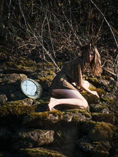 A contemplative nude figure sits gracefully among moss-covered volcanic rocks along Oregon's Floodplain Trail, bathed in dappled forest light. The subject's long auburn hair catches golden rays filtering through bare winter branches, while a circular mirror rests against the verdant stone formations. The intimate scene captures a moment of quiet introspection within the raw beauty of the Columbia River Gorge wilderness.