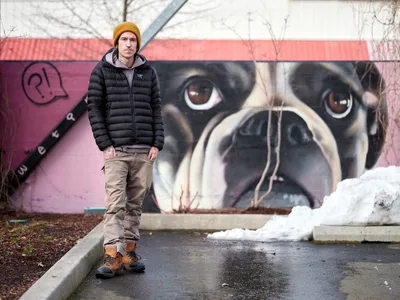 Painter and muralist Wetiko stands confidently before his completed dog portrait mural at the Clay Creative Building in Portland's Hosford-Abernethy neighborhood. The artist, wearing a golden mustard beanie and black puffer jacket, poses against the striking backdrop of his hyperrealistic canine artwork painted on pink-washed concrete walls. Winter's remnants are visible in patches of snow scattered across the wet pavement, while bare tree branches frame the scene under overcast Pacific Northwest skies.