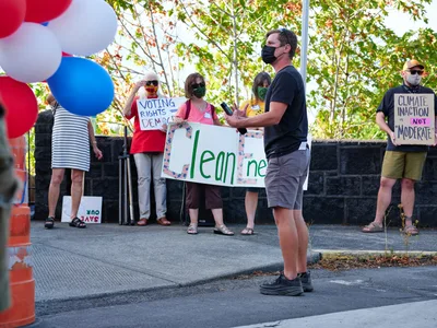 Organizer Michael Gaskill addresses a small gathering of environmental activists outside Representative Kurt Schrader's office on McLoughlin Promenade in Oregon City. Masked protesters hold handmade signs advocating for climate action, including messages about clean energy and democratic voting rights, while patriotic red, white, and blue balloons frame the scene. The afternoon light filters through autumn foliage, casting dappled shadows across the concrete plaza where citizens exercise their right to peaceful assembly.