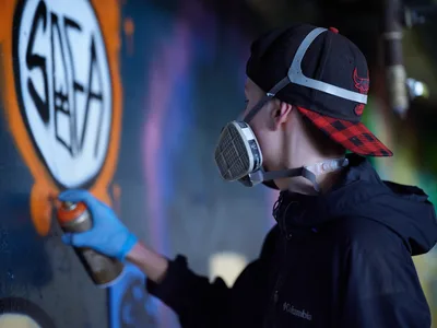 A street artist wearing a Chicago Bulls snapback and protective respirator carefully works on a vibrant mural within the industrial confines of Clay Creative's parking garage in Portland's Hosford-Abernethy neighborhood. The scene captures the intersection of urban art and workspace, as colored lighting bathes the concrete walls while the artist methodically applies paint to their canvas. The atmospheric garage setting transforms into a creative sanctuary, where spray paint cans and artistic vision merge under artificial illumination.
