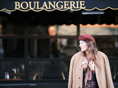 A woman in a burgundy beret and camel-colored coat stands contemplatively outside a traditional French boulangerie on Alberta Street in Portland, Oregon. The golden afternoon light catches her profile as she gazes thoughtfully to the side, her wavy brown hair framing her face beneath the classic French hat. The dark green storefront with ornate gold lettering creates an authentic European bakery atmosphere, while the shallow depth of field draws focus to her elegant silhouette against the blurred urban backdrop.