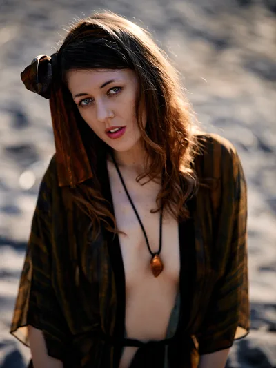 A young woman with flowing brown hair catches warm golden sunlight along the Floodplain Trail in Multnomah County, Oregon. She wears an olive military-style jacket over a simple top with an amber pendant necklace, her hand gracefully positioned near her hair as she gazes directly at the camera. The soft evening light creates a luminous halo effect around her silhouette against the blurred natural backdrop of the river valley landscape.