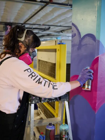 A focused artist wearing protective gear and a white 'PRIMTE' shirt applies vibrant purple spray paint to an emerging mural on a concrete wall. The industrial setting of Clay Creative's parking garage in Southeast Portland provides raw backdrop of exposed ceiling beams and yellow storage units. Her concentrated posture and precise movements capture the intimate process of transforming urban space into art, while spray cans rest on a wooden stepladder beside her.