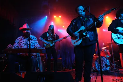 Barry Walker, Tobias Berblinger, and Victor Franco of Roselit Bone command the intimate stage at Dante's in Portland, their silhouettes carved against a wash of crimson and azure stage lights. Walker's cowboy hat catches the glow as he works the synthesizer, while Franco's leather jacket gleams under the dramatic lighting as he grips his white electric guitar. The atmospheric haze and moody lighting create an electric tension that defines Portland's underground music scene.
