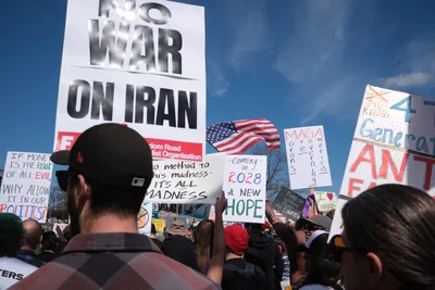 Protesters gather beneath a crystalline blue sky in Portland's historic Old Town district, their handmade signs creating a mosaic of dissent against potential military action. The crowd stretches into the distance along Southwest Ankeny Street, with prominent placards declaring "NO WAR ON IRAN" and references to 2028 hope rising above a sea of baseball caps and winter jackets. An American flag waves paradoxically among the anti-war messages, while demonstrators of various ages stand shoulder-to-shoulder in peaceful assembly.